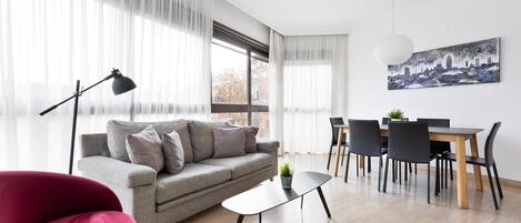 Una sala moderna con un sofá gris, un sillón rojo, una mesa de comedor con sillas y un cuadro de un paisaje urbano en la pared.