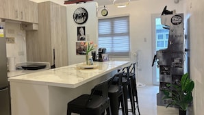 A modern kitchen with a marble countertop, bar stools, a ship decoration, and a wall-mounted fan. - Cozy Loft Condo in Tanza With Arcade (Tanza)