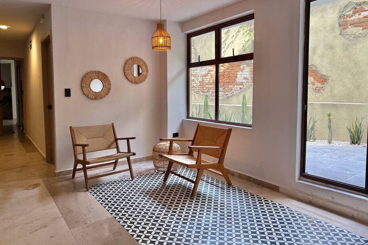 Una stanza con due sedie di legno, una panca intrecciata, un tappeto decorato e un'ampia finestra che dà su un muro di mattoni e piante.