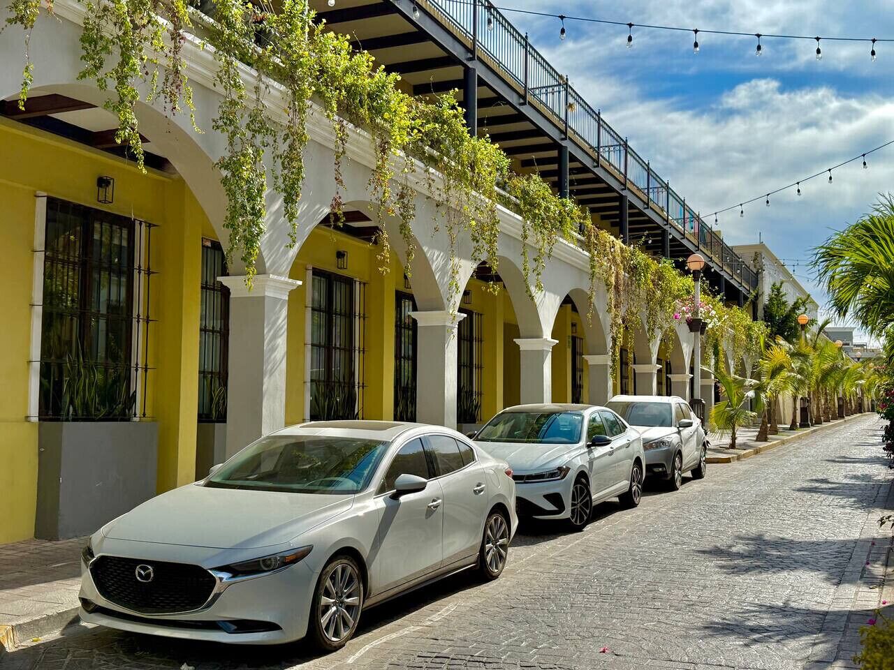 A street with parked cars and a building with arches and hanging plants.