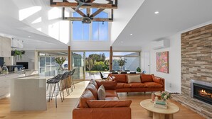 Un espacio habitable amplio con un sofá grande, una mesa de centro y una chimenea. Se ve un área de cocina con una barra y un comedor a través de amplias ventanas.