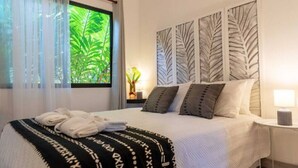 A neatly made bed with a patterned blanket, a window with sheer curtains, and a wall with leaf-patterned artwork. - La Prometida - House Marea (Puerto Viejo)