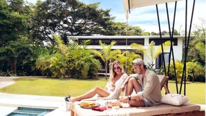 Una pareja descansando junto a una piscina en un sillón con vistas a vegetación exuberante y un edificio moderno al fondo.
