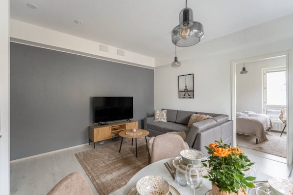 Una sala de estar moderna con televisor de pantalla plana, un sofá gris y una zona de comedor con una mesa redonda para dos.