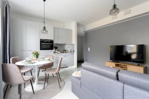 Una sala de estar moderna con un sofá gris, una mesa de comedor con sillas, una zona de cocina y un televisor de pantalla plana montado en la pared.