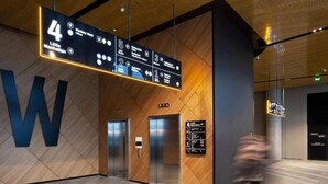 Un espace moderne avec des ascenseurs, comprenant un grand panneau en forme de « W », un panneau d’affichage numérique indiquant les différents étages, et deux ascenseurs.