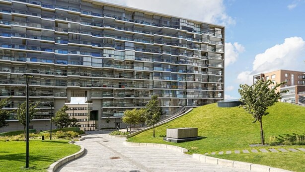 Un immeuble résidentiel à plusieurs étages, doté de balcons, d’une cour aménagée et d’une petite colline avec un sentier menant à l’entrée.