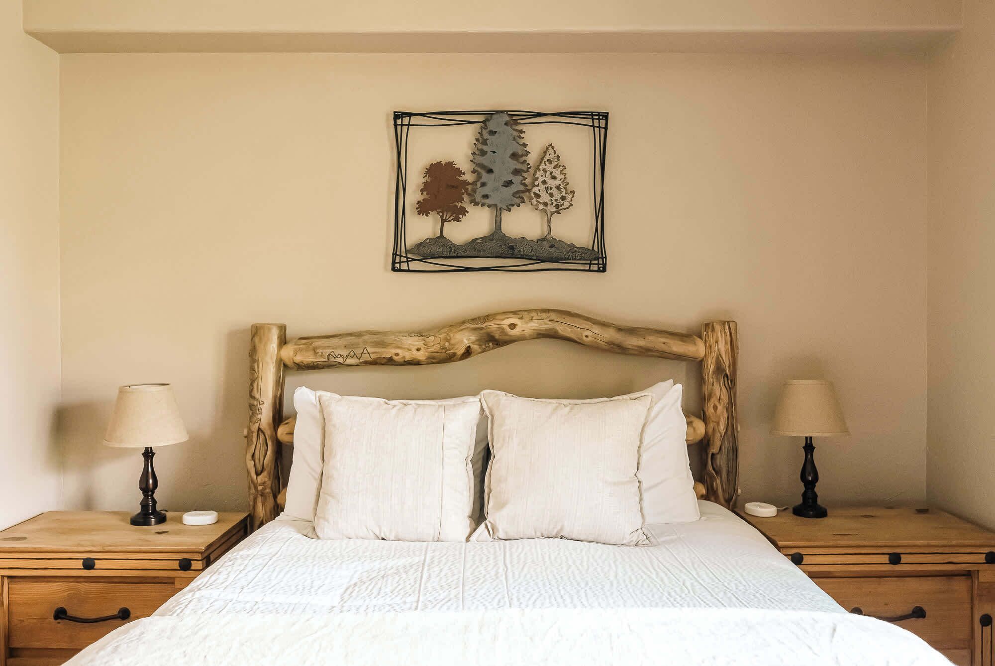 A bedroom with a wooden headboard, a bed with white bedding, two bedside tables with lamps, and a wall-mounted artwork of trees.