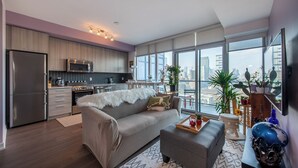 Un apartamento moderno con cocina, comedor y sala de estar. Cuenta con un amplio ventanal con vista a la ciudad, un sofá gris y una mesa de comedor de madera.