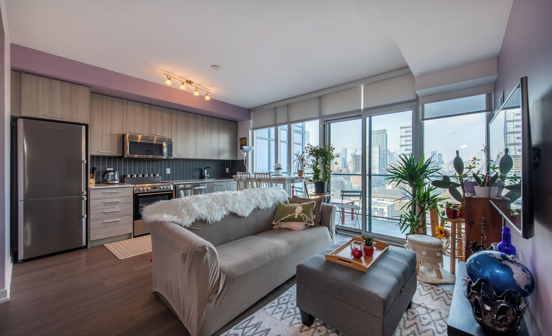 A modern apartment with a kitchen, dining area, and living room. It features a large window with a city view, a grey sofa, and a wooden dining table.