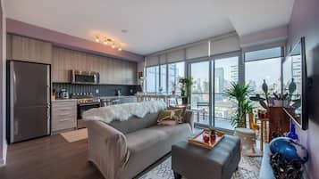 A modern apartment with a kitchen, dining area, and living room. It features a large window with a city view, a grey sofa, and a wooden dining table.