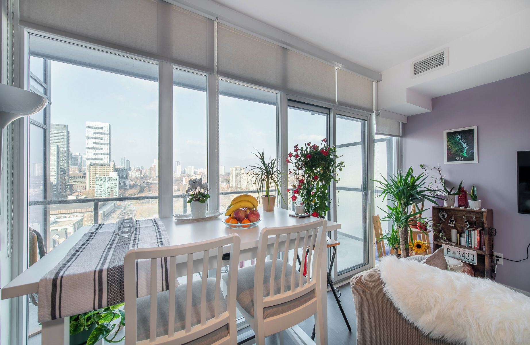 A modern dining area with a city view, a dining table set with a placemat, chairs, and a bowl of fruit.