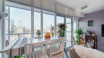A modern dining area with a city view, a dining table set with a placemat, chairs, and a bowl of fruit.