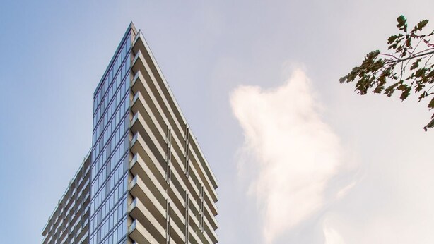 Un edificio moderno de gran altura con fachada de vidrio, varios balcones y un estacionamiento al frente.