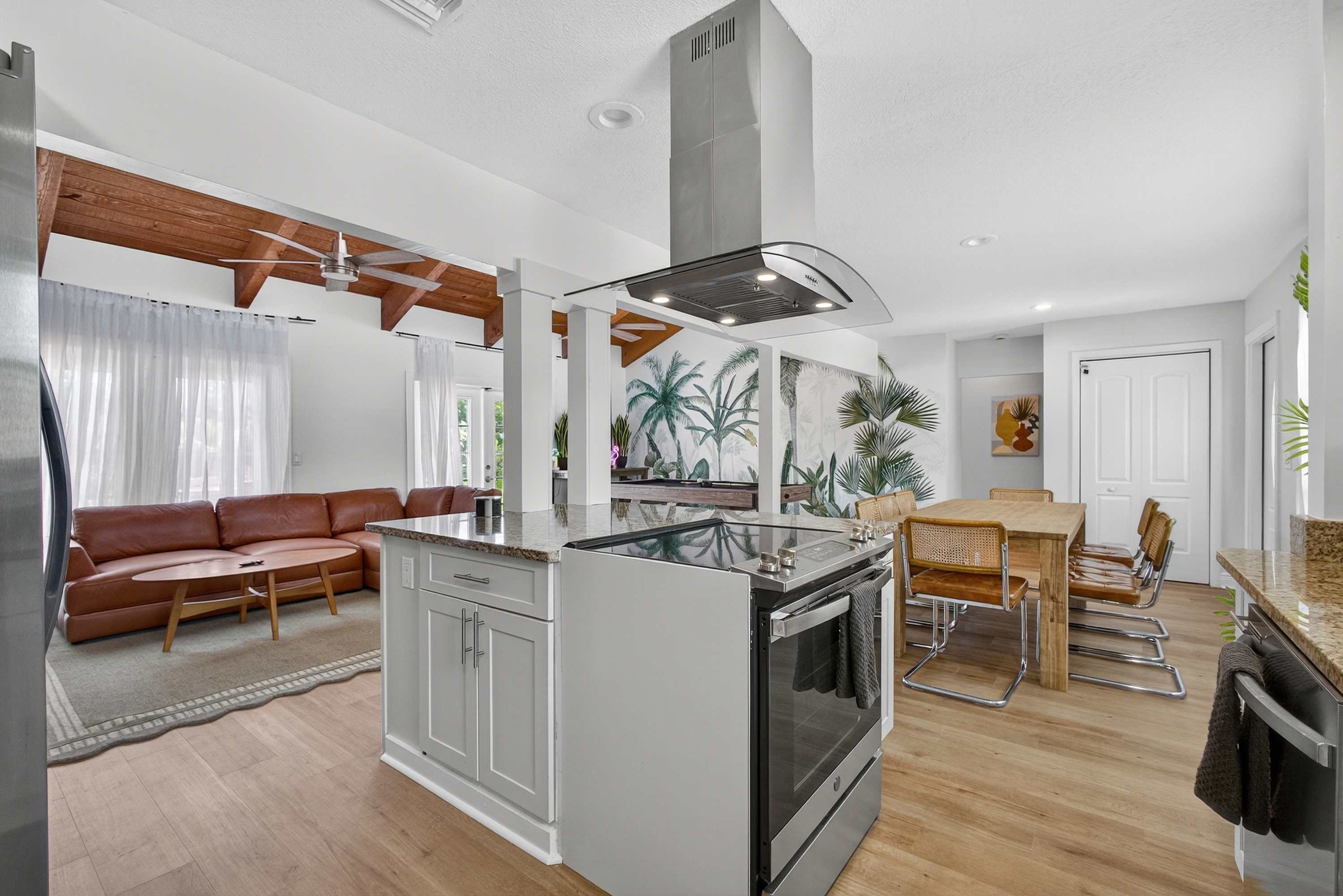 A modern kitchen with a central island, a dining area with a wooden table and chairs, and a living area with a brown leather sofa and a glass-top coffee table.