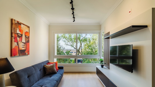 A modern living room with a dark blue sofa, a round white table, a mounted flat-screen TV, and a large window offering a view of trees and buildings. - Cozy and Well-located Apartment in POA - Rfa303 (Porto Alegre)