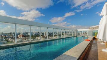 Piscina infinita no terraço, com vista para a cidade, cercada por grades de vidro e deck de madeira.