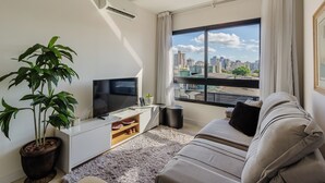 A modern living room with a large window, a flat-screen TV, a sofa, a potted plant, and a small side table. - Lindo Apto em Cond. c\/ Piscina e Garagem RPF0605 (Porto Alegre)