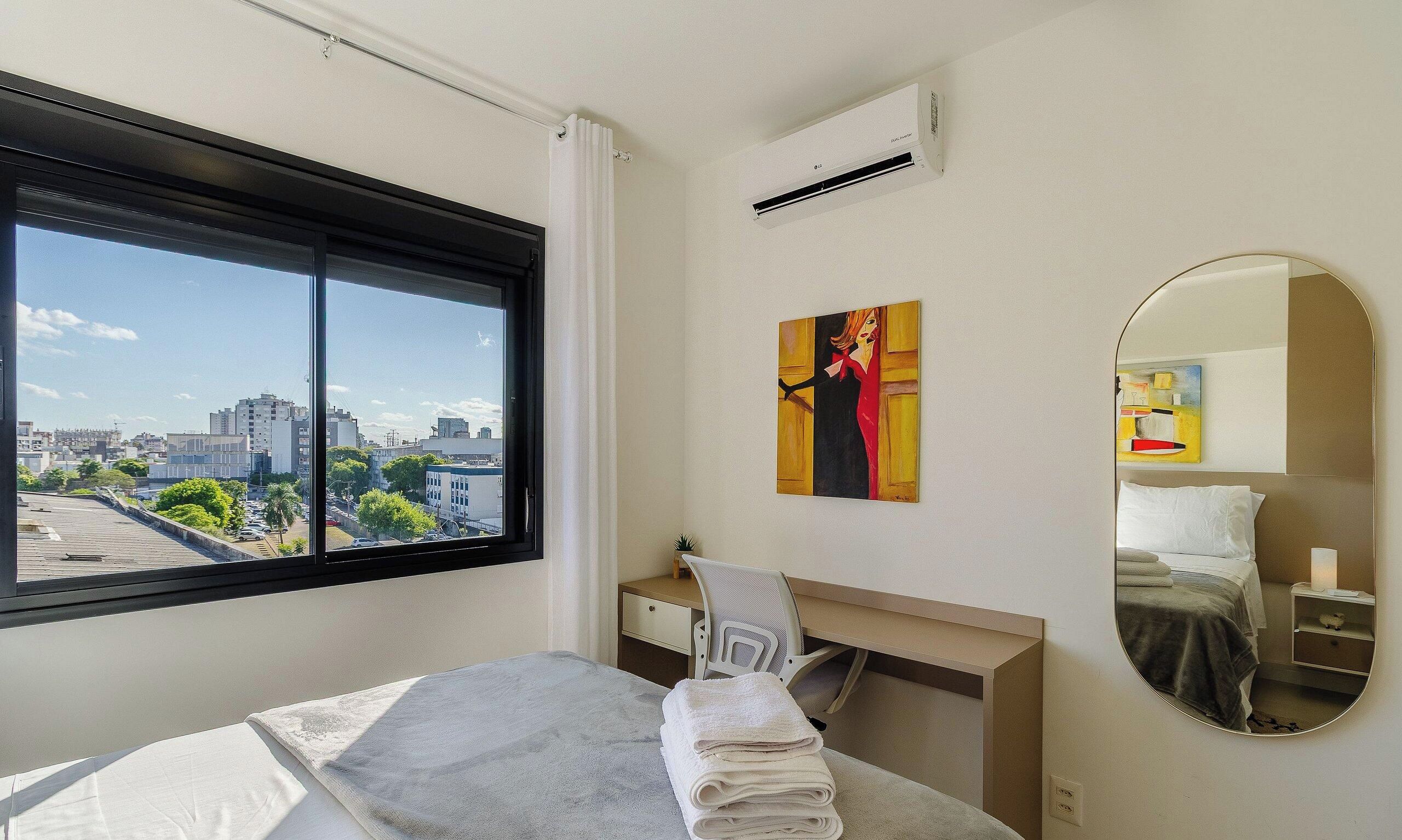 Quarto de hotel com uma janela grande, uma cama, uma escrivaninha, uma cadeira, um quadro, um espelho e um ar condicionado.