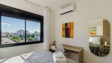 Quarto de hotel com uma janela grande, uma cama, uma escrivaninha, uma cadeira, um quadro, um espelho e um ar condicionado.