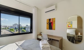A hotel room with a large window, a bed, a desk, a chair, a painting, a mirror, and an air conditioning unit. - Lindo Apto em Cond. c\/ Piscina e Garagem RPF0605 (Porto Alegre)