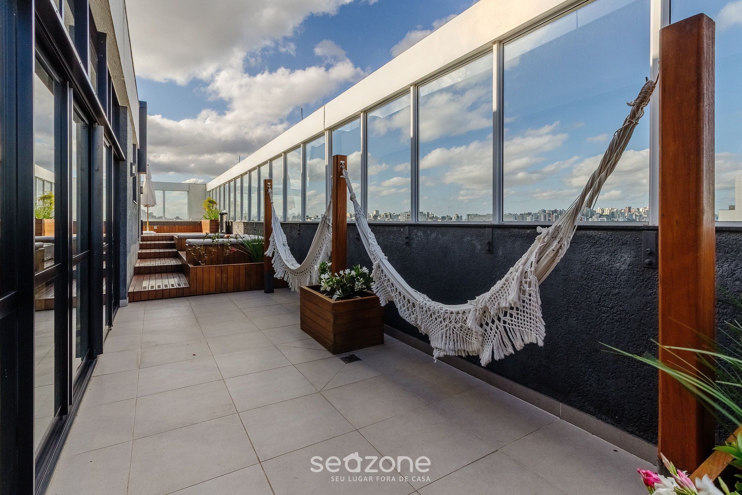 Terraço no telhado com grandes janelas, uma rede e plantas em vasos.