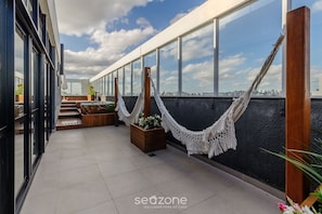 A rooftop terrace with large windows, a hammock, and potted plants. - Lindo Apto em Cond. c\/ Piscina e Garagem RPF0605 (Porto Alegre)