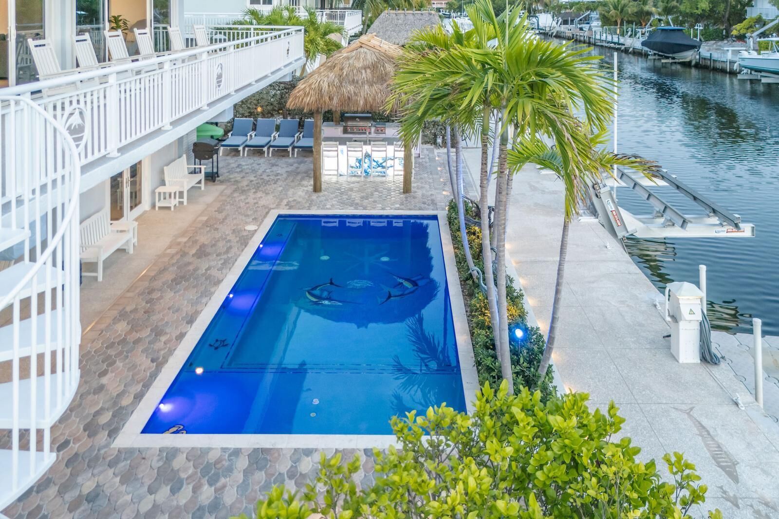 A swimming pool with lounge chairs and a thatched roof structure, surrounded by palm trees and a marina.