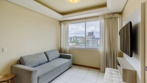 A living room with a grey sofa, a small wooden stool, a white sofa, a TV mounted on the wall, and a large window with curtains. - Elegant Apartment for Couples in Poa-rs Urb1110 (Porto Alegre)