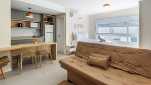 Une pièce moderne et bien éclairée, dotée d’une kitchenette, d’un coin repas avec une table et des chaises en bois, d’une grande fenêtre avec des stores et d’un canapé marron agrémenté d’un coussin.