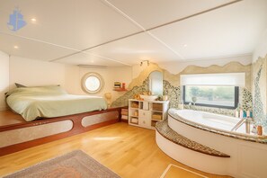 Een slaapkamer met een bed, een rond raam, een ingebouwde kaptafel met wastafel en een stenen muur.
