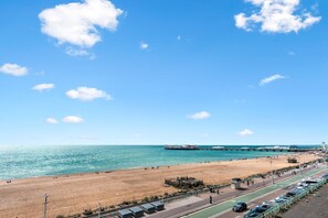 A beach with a pier, a parking area, and a road with cars.