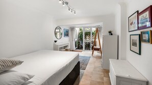 A modern bedroom with a bed, a desk, and a chair. There is a balcony with a view of greenery.
