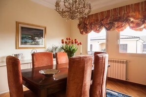 A dining room with a wooden table, six chairs, a chandelier, a painting, and a window with curtains.