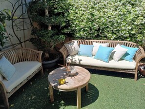 A wooden outdoor seating area with a round table and two wicker chairs, surrounded by greenery and a flowering bush.