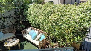 An outdoor seating area with a wooden sofa and a small round table, surrounded by lush greenery and white flowering bushes.