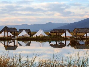 A series of thatched-roof tents and white tents by a calm body of water with mountains in the background. - ENDEMICA GLAMP RESORT (Cuatro Cienegas)