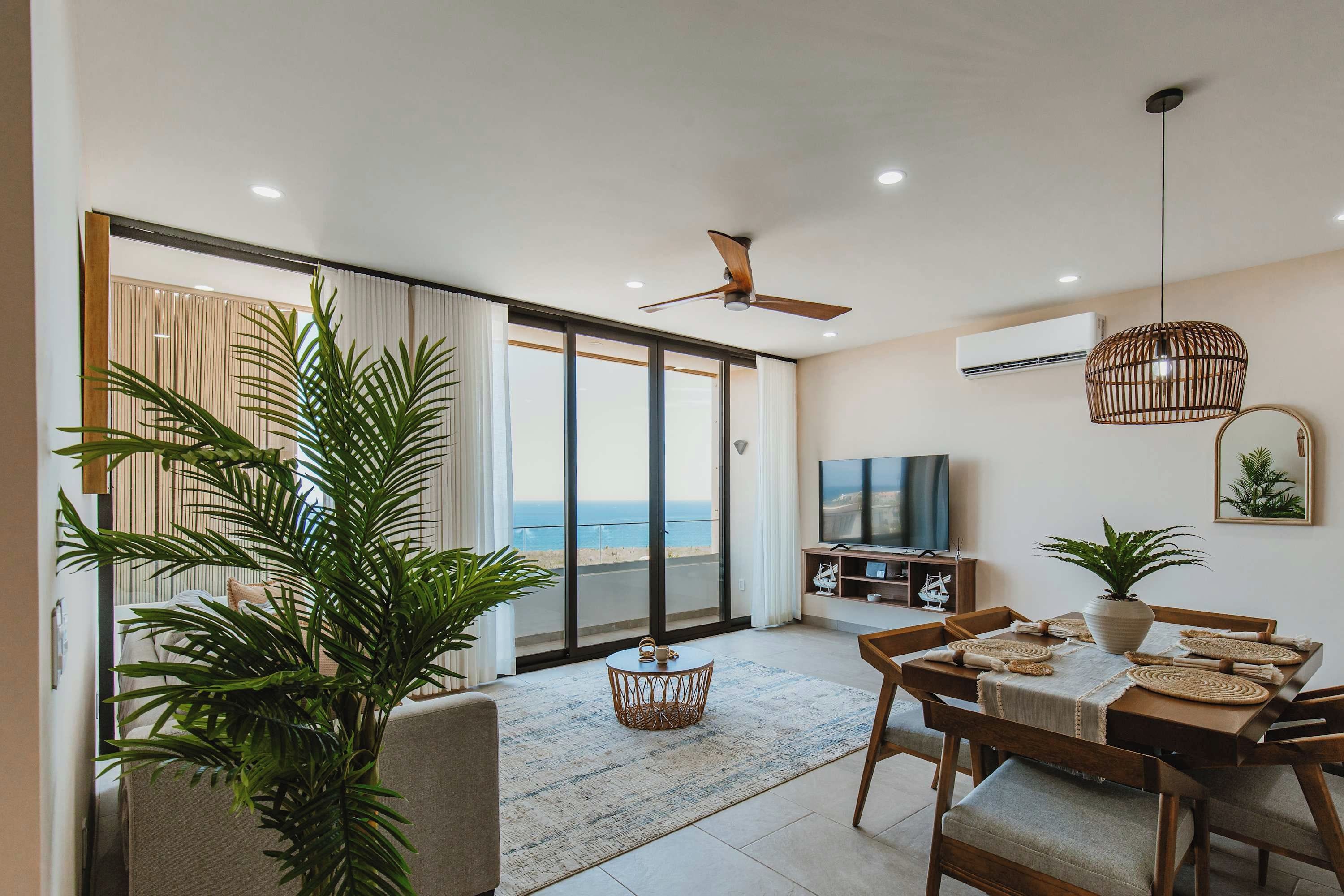 A spacious living area with a large sliding glass door leading to a balcony, a ceiling fan, a TV, and a dining table set with placemats.