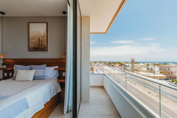 A bedroom with a large bed, a bedside table, a nightstand, a window, and a view of a road and buildings.