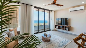 A modern living room with a large TV, a ceiling fan, and a view of the ocean through large sliding doors. - Arch View Condo Cabo San Lucas • 4th Floor Rooftop Pool | 403 (Cabo San Lucas)