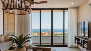 A modern living room with a large window, a ceiling fan, a potted plant, and a flat-screen TV. - Arch View Condo Cabo San Lucas • 4th Floor Rooftop Pool | 403 (Cabo San Lucas)