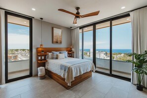 A bedroom with a large bed, a ceiling fan, a view of the city, and a balcony with a plant.