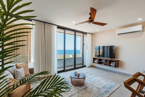 A modern living room with a large TV, a ceiling fan, and a view of the ocean through large sliding doors.
