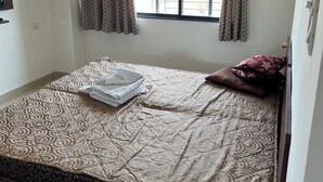 Ein Einzelbett mit gemusterten Betttextilien, ein Fenster mit Vorhängen und ein an der Wand befestigter Fernseher.