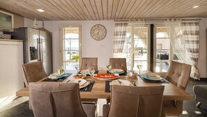 A dining area with a wooden table set for four, surrounded by cushioned chairs. A wall clock and a window with curtains are visible. - Villa Ventina with Pool (Senj)