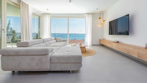 Un salon moderne doté d’un grand canapé d’angle, d’un téléviseur fixé au mur et d’un balcon offrant une vue sur la mer.