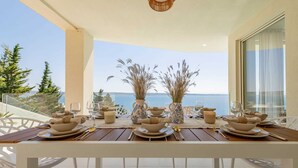 Une table à manger pour quatre personnes, avec des chaises blanches, est placée sur un balcon d’où l’on peut admirer la mer et les arbres.