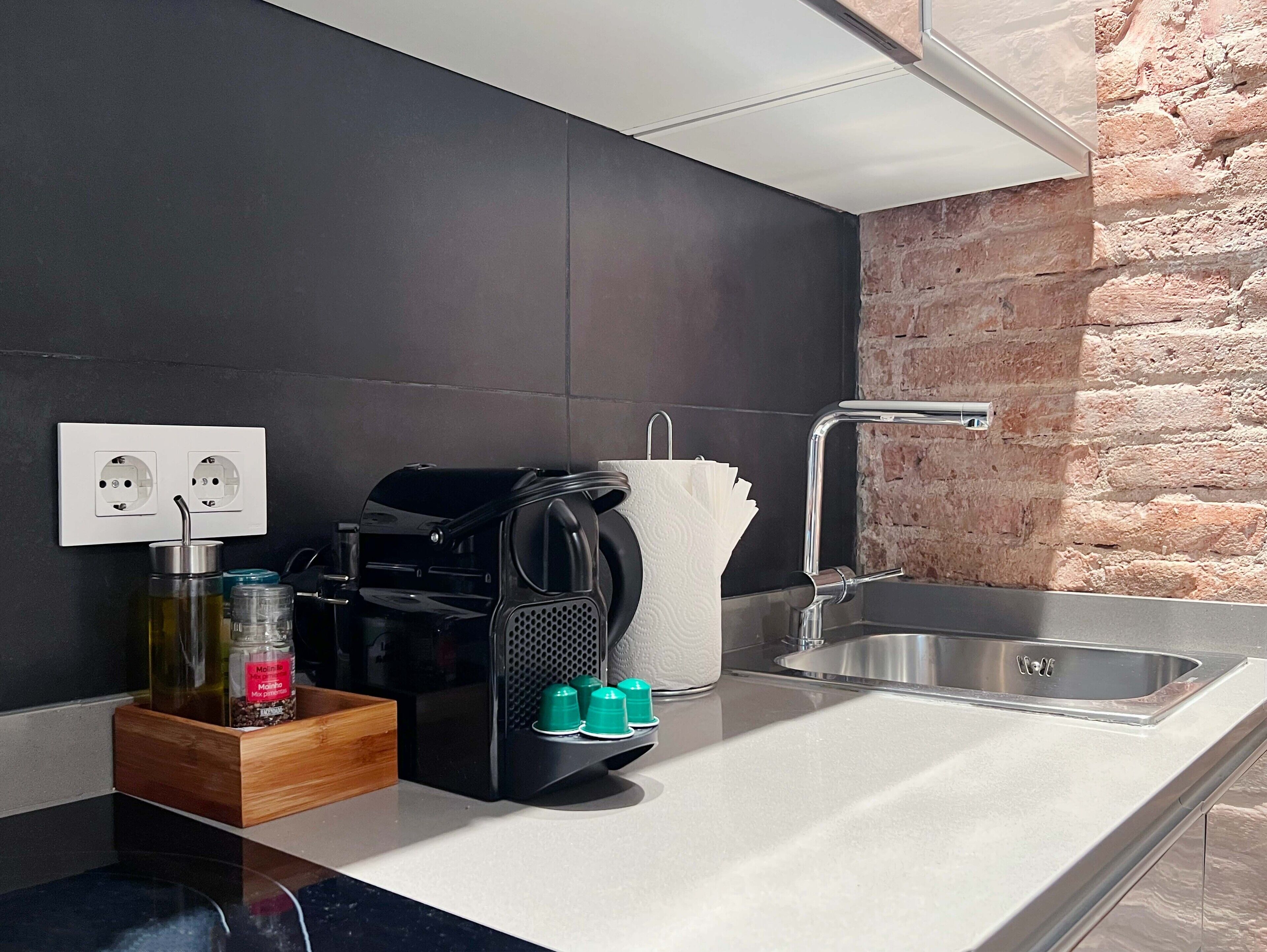 A modern kitchen with a black countertop, a built-in oven, a coffee machine, and a sink with a brick wall backsplash.