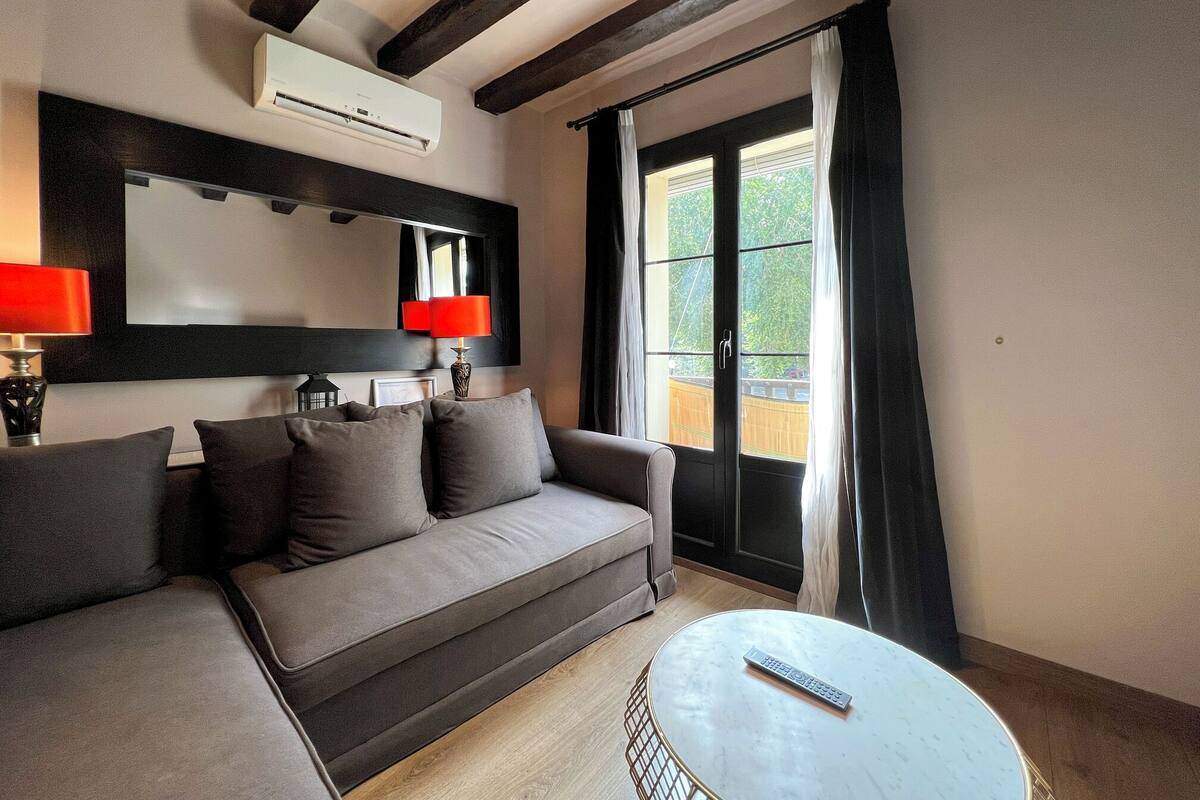A modern living room with a sofa, a round coffee table, a remote control, a red lamp, and a large window with curtains.