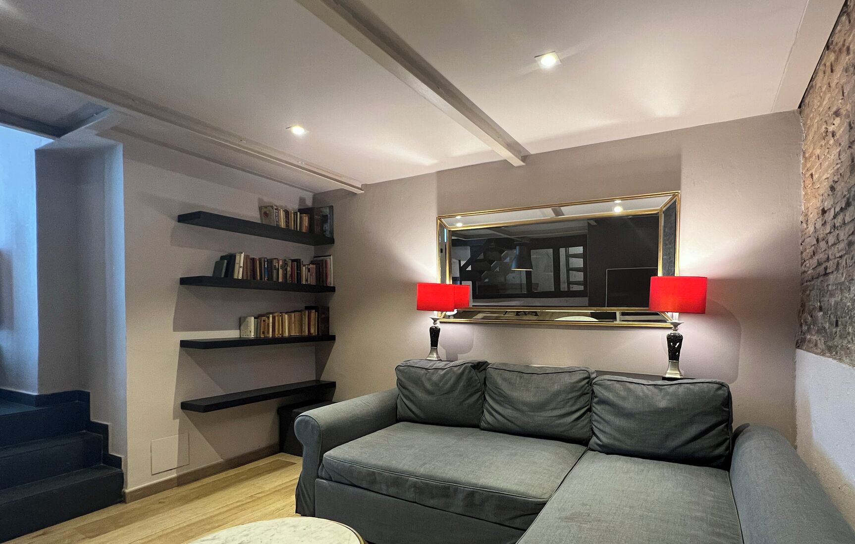A modern living room with a grey sofa, a bookshelf, a large mirror, and two red lamps.